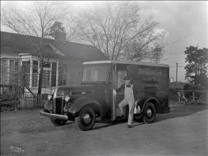 A white man wearing overalls and a black cap stands in the door of an old fashioned delivery truck in front of a one story house and some trees. The man is holding glass milk bottles. 